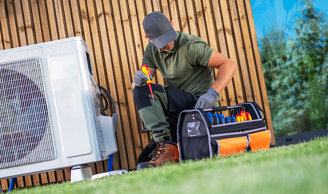 A technician repairs an air conditioning unit outside, meticulously organizing tools in a bag while seated on the grass.